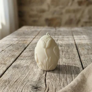 Vela decorativa en forma de huevo con relieve de flor en color blanco sobre mesa rústica de madera