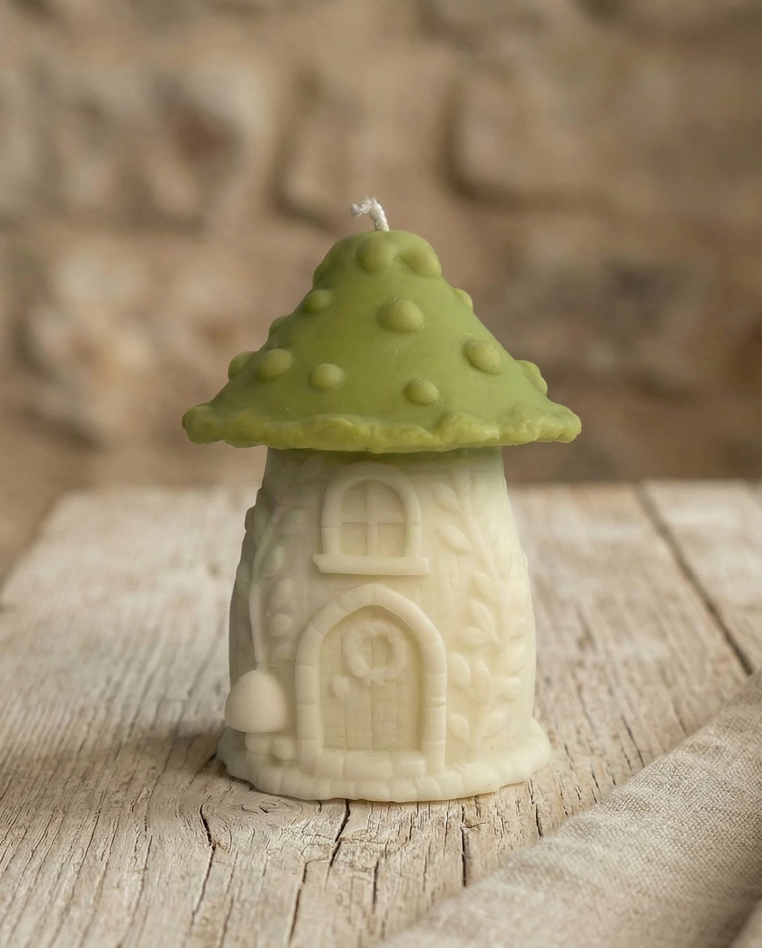 Vela decorativa en forma de seta casita de gnomo con tejado verde sobre mesa de madera rústica