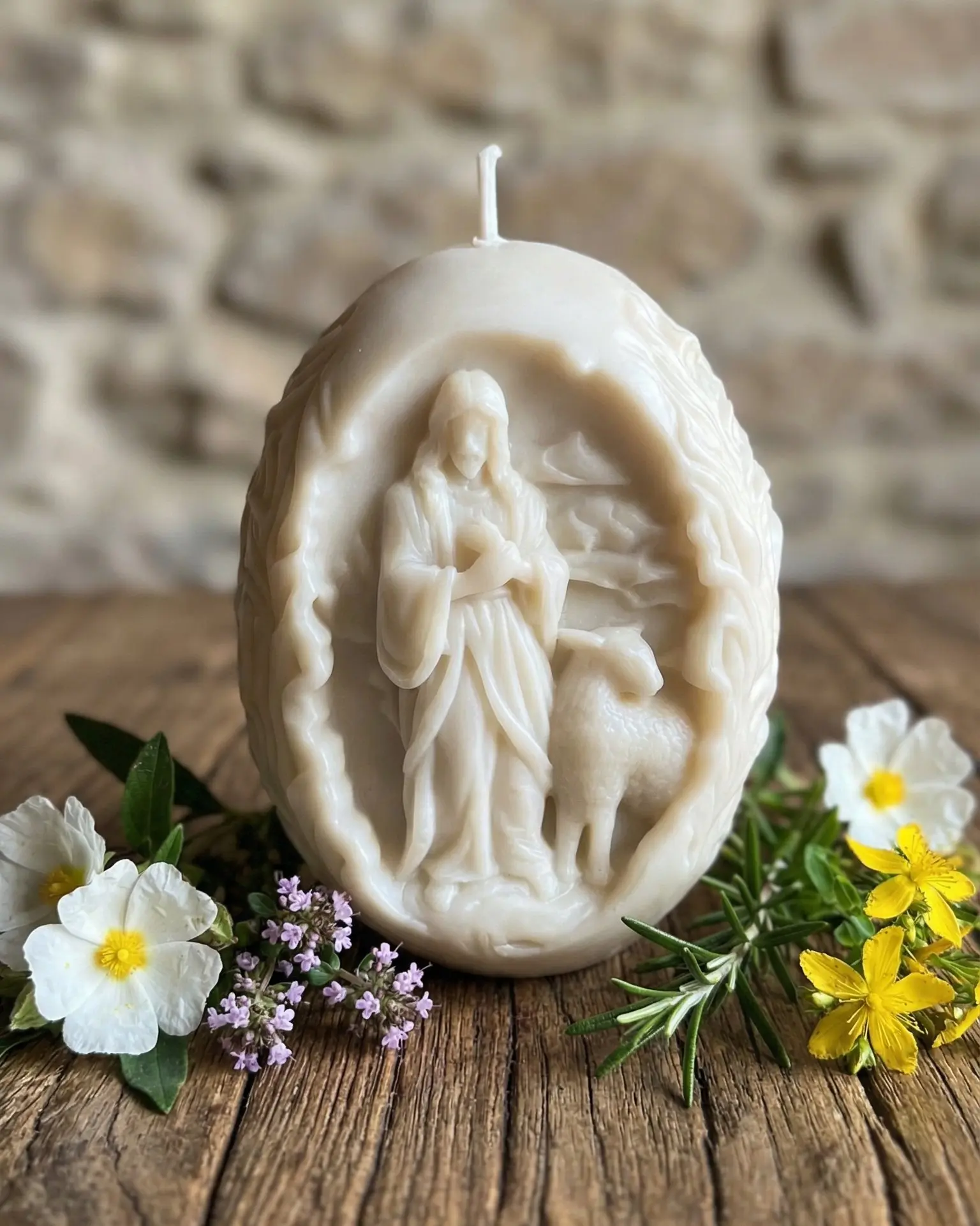 Vela religiosa Cordero de Dios de soja natural con flores sobre mesa de madera rústica